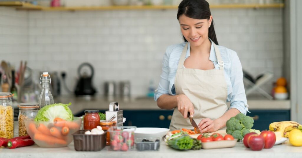 Woman chopping vegetables in a bright kitchen surrounded by colorful fresh produce, representing nourishing, healing meals made with real food.
