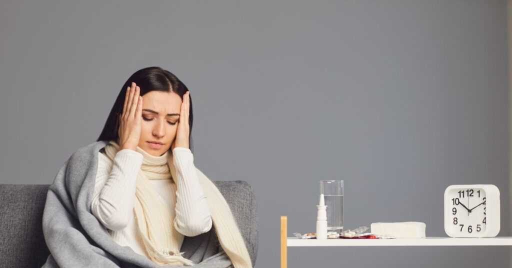 Woman wrapped in a blanket holding her head in discomfort, sitting beside a table with medications and a clock, symbolizing the struggle of persistent symptoms and the search for answers.