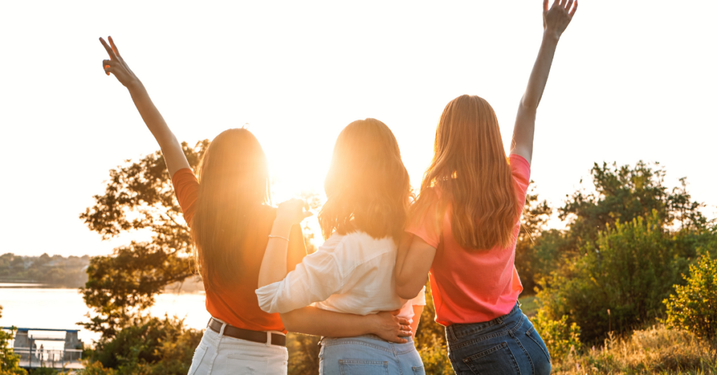 Three women standing together with arms raised at sunset.